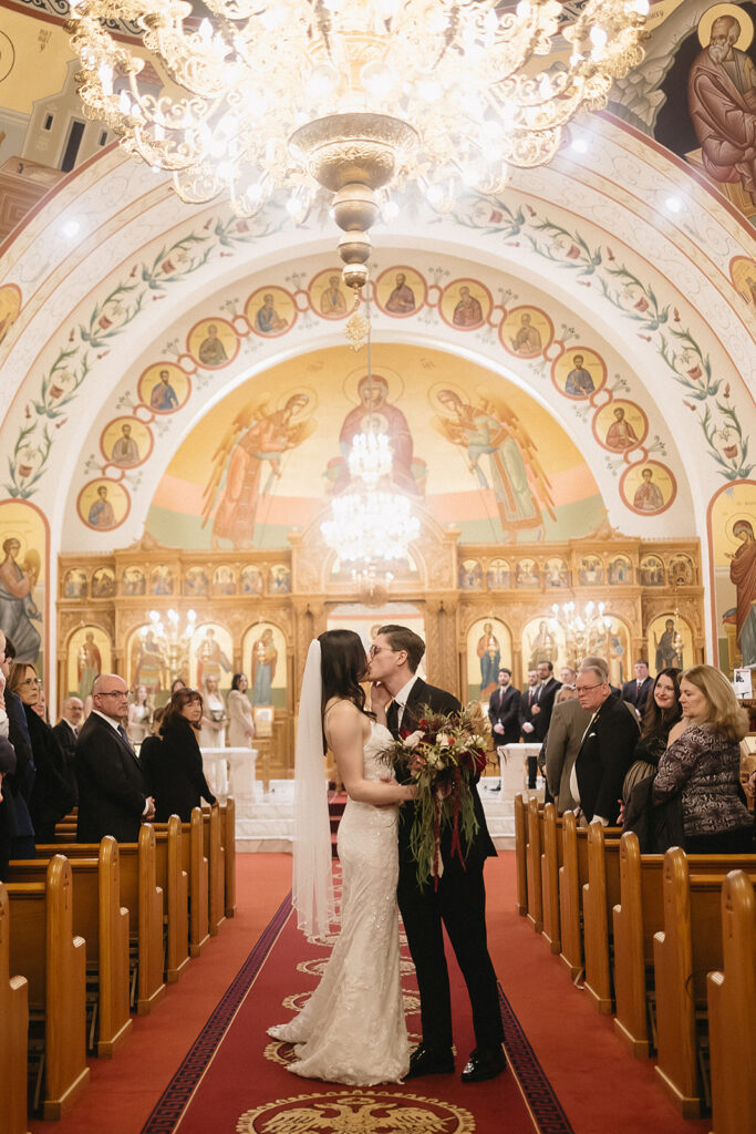 A bride and groom kiss in the aisle of a beautifully decorated church with ornate chandeliers, religious icons, and guests seated in pews on either side, celebrating their wedding ceremony.