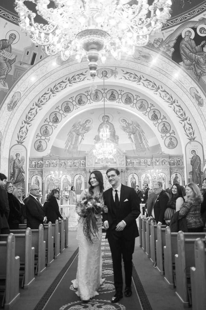 A bride and groom walk down the aisle of an ornate church, smiling as guests seated on both sides watch. The setting features chandeliers, arches, and religious iconography. The image is in black and white.