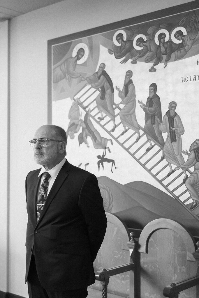 A man in a suit stands in front of a religious mural depicting figures climbing a ladder toward saints or angels, with others falling off. The scene is indoors and captured in black and white.