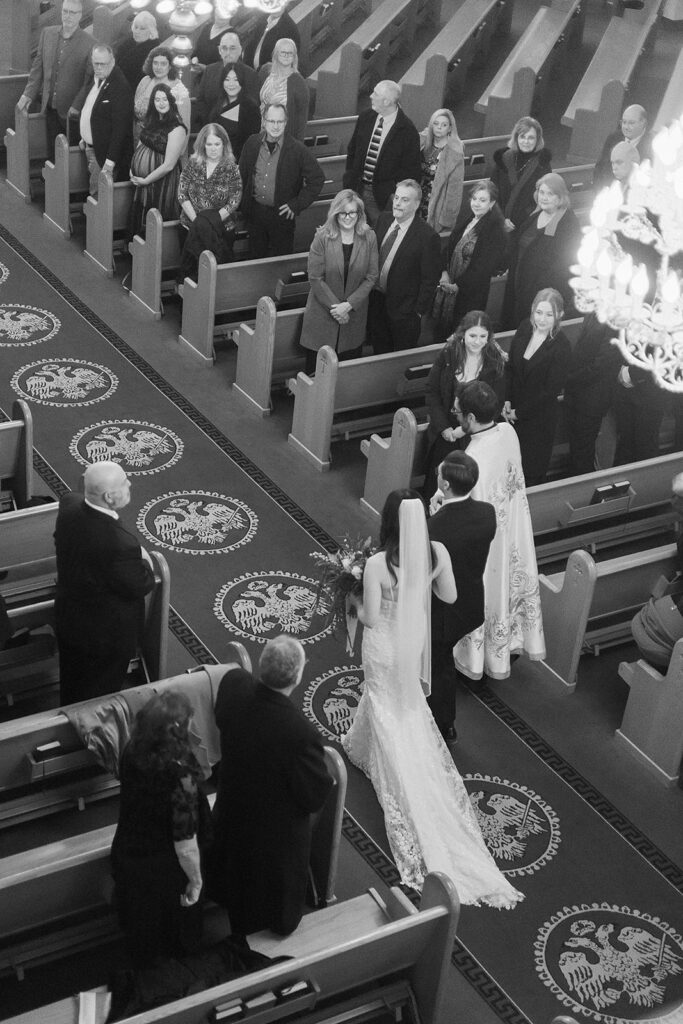 A black-and-white photo of a bride and groom at the altar in a church, surrounded by standing guests in pews, as a wedding ceremony takes place. The guests are watching the couple, and chandeliers hang above.