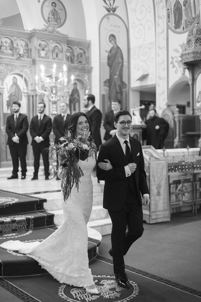 A bride and groom walk arm in arm down the aisle in an ornately decorated church. The bride wears a long white gown and holds a bouquet, while the groom is in a dark suit. People stand in the background, watching.