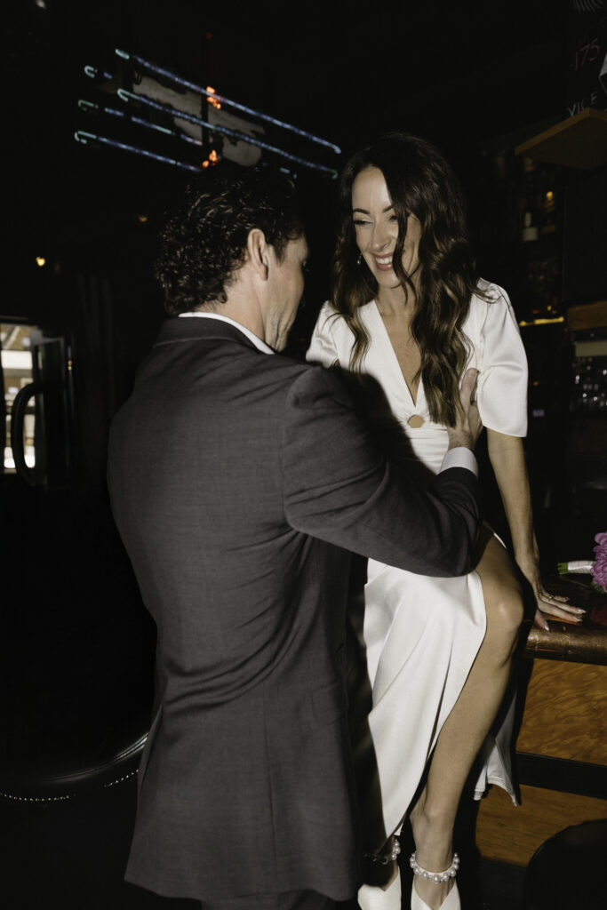 A man in a suit stands close to a smiling woman in a white dress sitting on a bar counter, her hand resting on his shoulder, in a dimly lit bar or lounge setting.