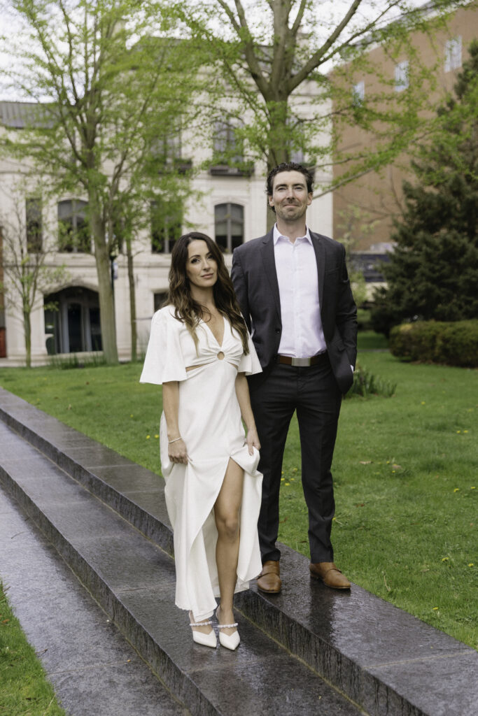 A woman in a white dress and a man in a dark suit stand side by side outdoors on stone steps, surrounded by green grass, trees, and buildings in the background.