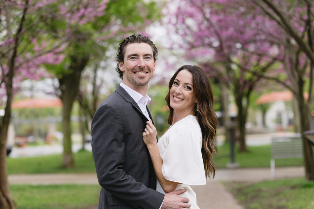 A smiling couple poses outdoors on a pathway lined with blossoming trees. The man wears a dark suit and the woman wears a white dress. Pink flowers and greenery fill the background, creating a springtime atmosphere.