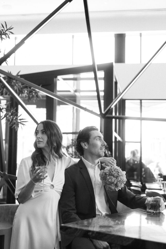 A woman in a light dress holds a glass of wine and sits beside a man in a suit holding flowers and a drink at a modern, indoor setting with large windows and geometric decor. The mood is relaxed and elegant.