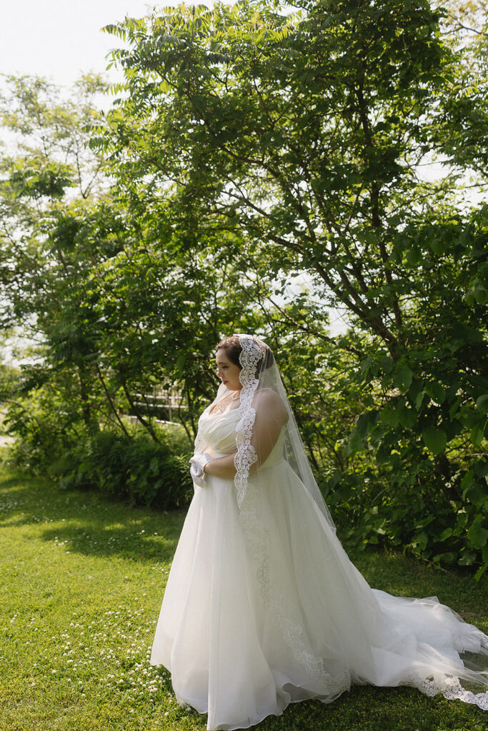 A bride in a white gown and lace veil stands on green grass surrounded by lush trees, looking down and holding her hands together in front of her. Sunlight filters through the leaves, creating a serene, natural scene.