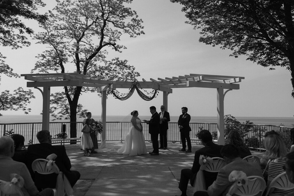 A wedding ceremony takes place outdoors under a white pergola, with the couple, officiant, and two attendants standing. Guests are seated, and trees frame the view of a large body of water in the background.