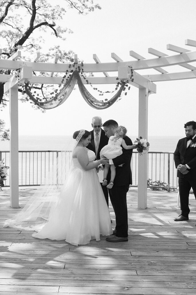 A bride and groom stand facing each other during an outdoor wedding ceremony under a decorated pergola, holding a small child. An officiant and another man stand nearby. The scene overlooks water in the background.