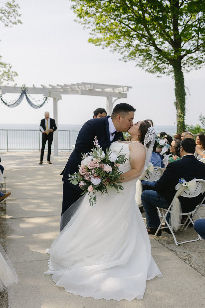 A bride and groom share a kiss during their outdoor wedding ceremony, surrounded by guests seated in white chairs, with trees and a view of water in the background. The bride holds a bouquet of pink and white flowers.