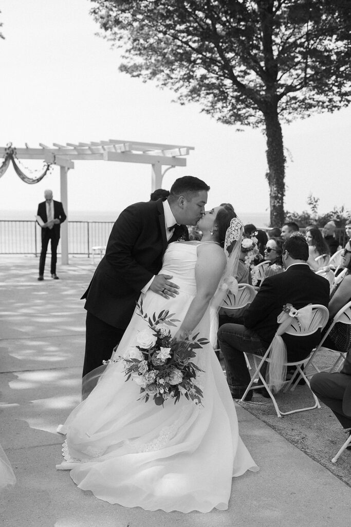 A groom kisses his bride as they walk down the aisle outdoors, surrounded by seated guests. The scene is shaded by trees with a decorated pergola and a man standing in the background. The image is in black and white.