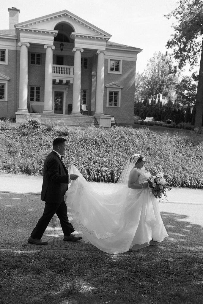 A groom follows a bride, holding her dress train as they walk outside. The bride holds a bouquet and wears a veil. In the background is a large elegant building with columns and steps. The photo is in black and white.