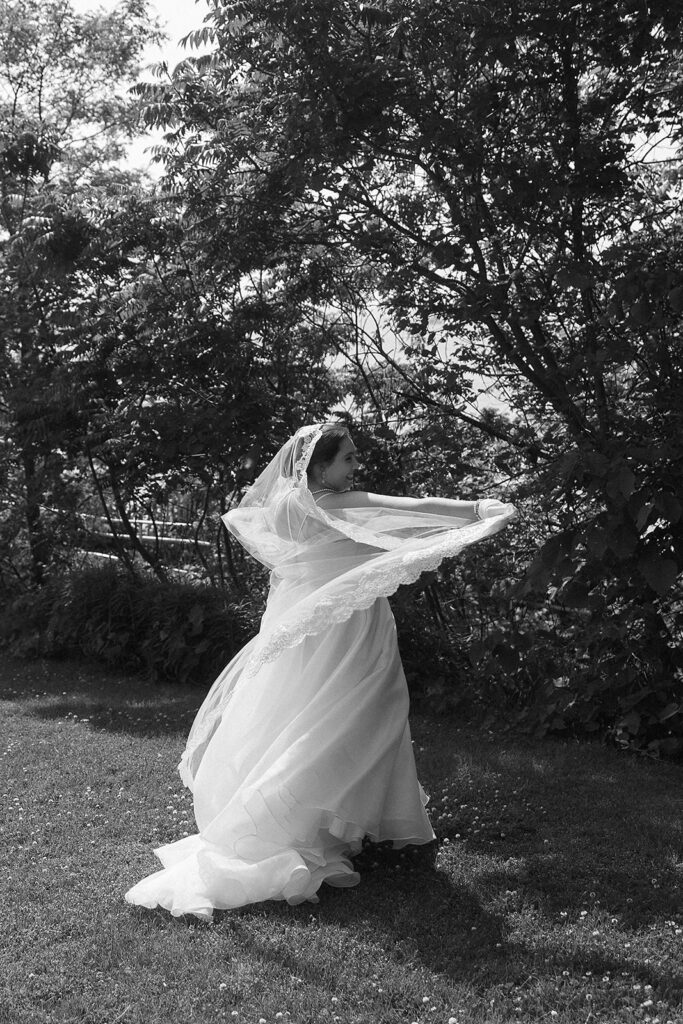 A bride in a flowing gown and veil stands on grass, smiling and holding her veil outstretched with trees and shrubs in the background. The photo is in black and white.