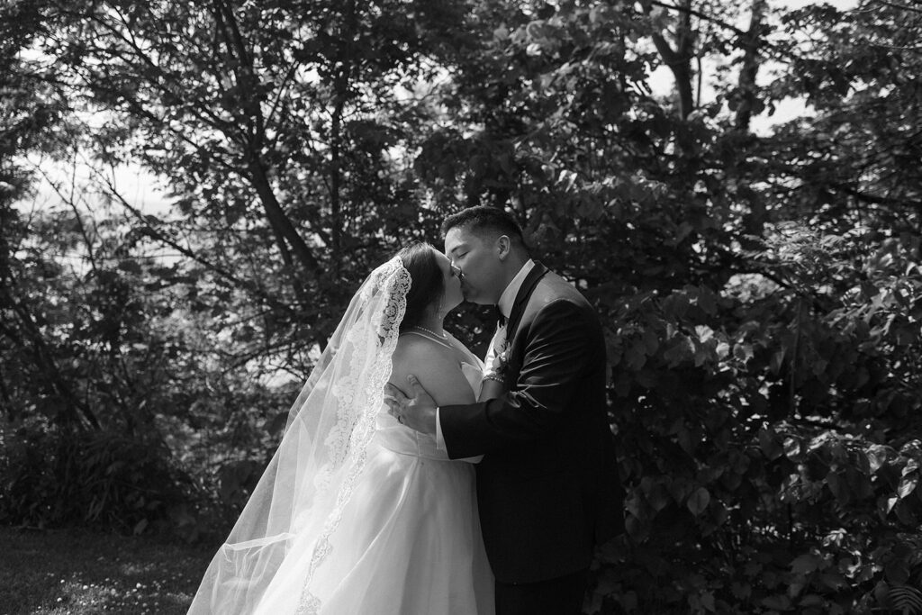 A bride and groom share a kiss outdoors, surrounded by trees. The bride wears a veil and a white dress, while the groom is in a suit. Sunlight filters through the foliage, creating a romantic atmosphere.