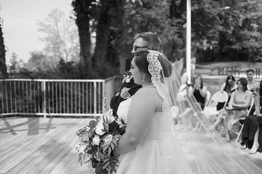 A bride in a lace veil holds a bouquet and stands beside an older man in a suit, likely her father, during an outdoor wedding ceremony. Guests are seated and watching in the background.