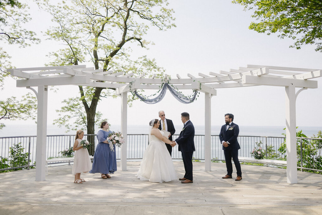 A bride and groom stand under a white pergola during an outdoor wedding ceremony by the water, holding hands with an officiant behind them. Bridesmaids and a groomsman stand on either side, surrounded by greenery.