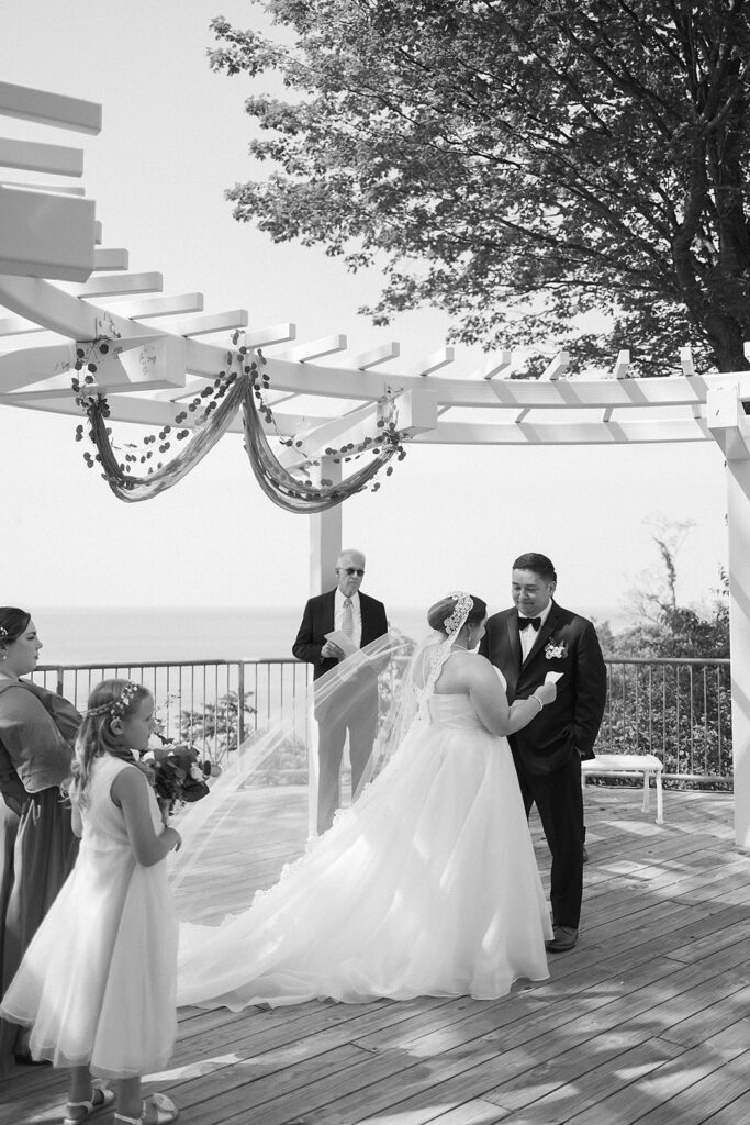 A bride and groom stand facing each other during an outdoor wedding ceremony under a decorated pergola, with an officiant and two flower girls nearby. The scene overlooks water and trees in the background.