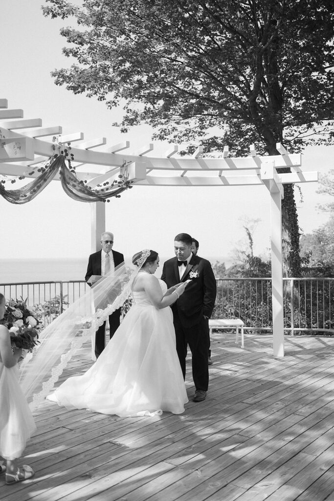 A bride and groom stand facing each other on a wooden deck under a pergola, holding hands during their outdoor wedding ceremony. A flower girl and an officiant are nearby. The setting overlooks the water, with trees in the background.
