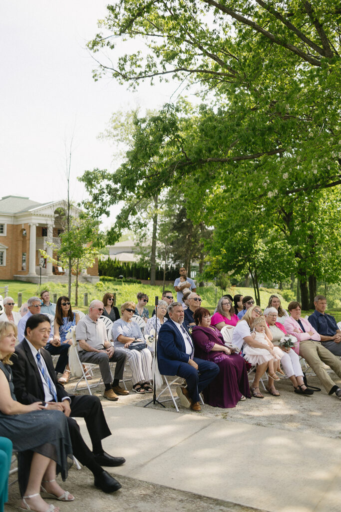 A group of people sit outdoors on folding chairs, attentively watching an event. They are surrounded by green trees and a large brick building is visible in the background. The atmosphere appears calm and formal.