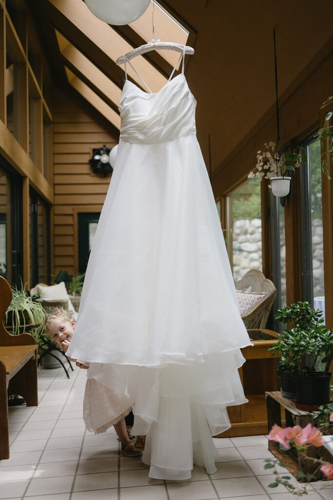 A white wedding dress hangs indoors, and a young child in a light dress peeks out playfully from underneath the gown. The setting is bright, with wooden walls, windows, and various potted plants.