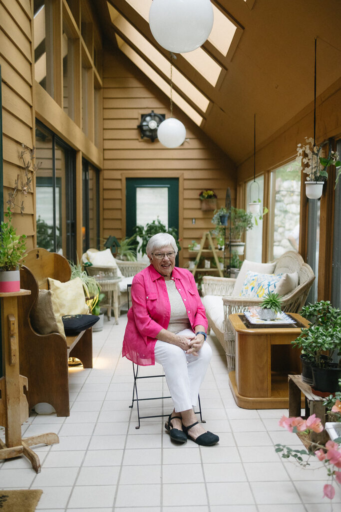 An elderly woman with short gray hair, wearing a bright pink cardigan and white pants, sits on a chair in a sunlit, plant-filled indoor patio with large windows and wooden walls.