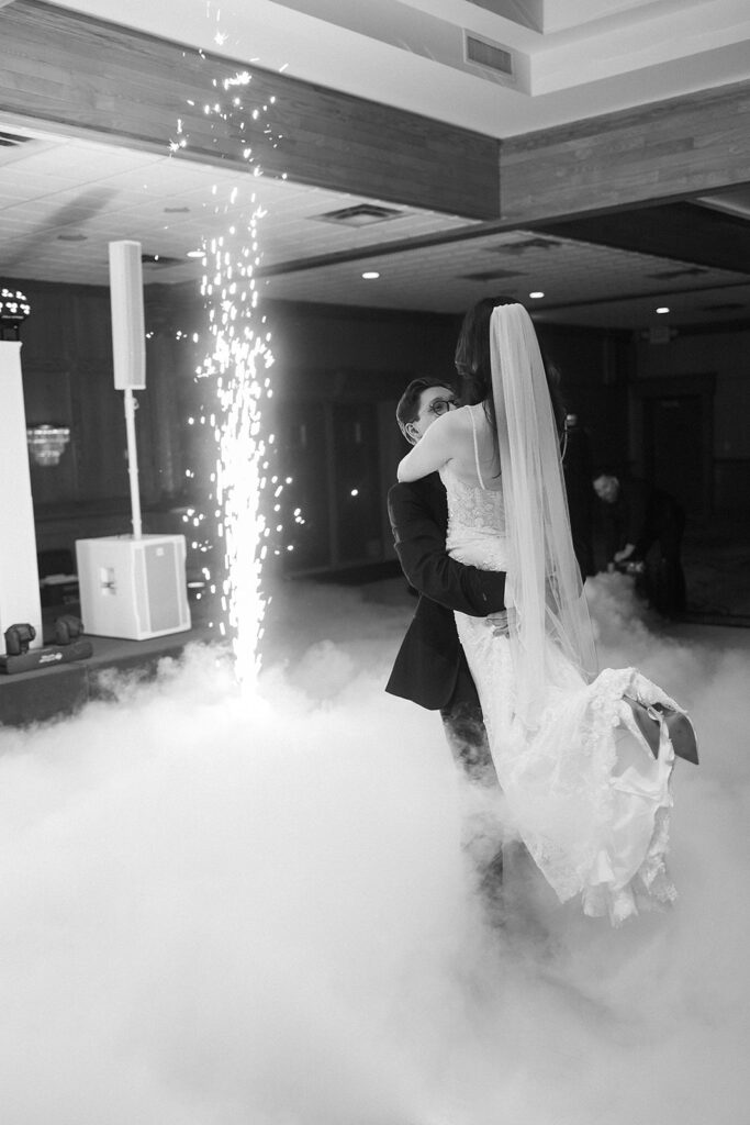 A groom lifts his bride in a dance surrounded by fog and indoor fireworks, in a decorated indoor venue. The bride wears a long veil and lace dress; the moment is joyful and romantic.