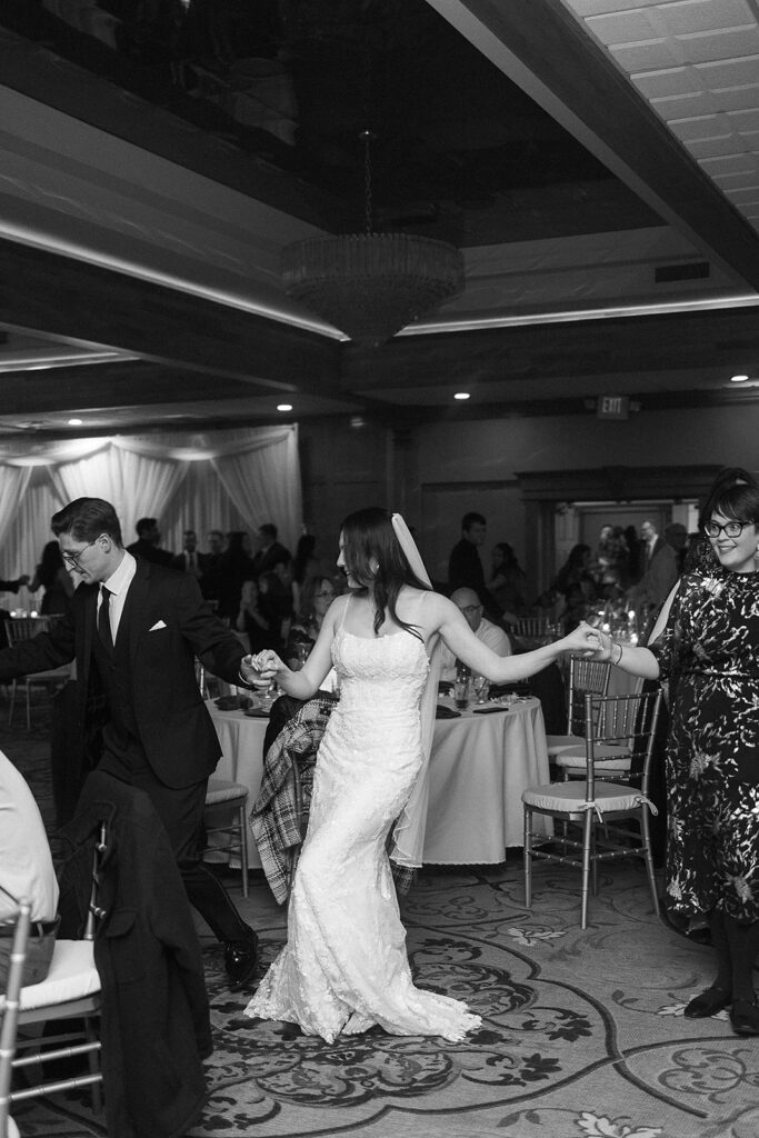A bride in a white dress and veil holds hands with a groom in a suit and a guest, leading a lively dance in a decorated reception hall with tables and guests in the background.