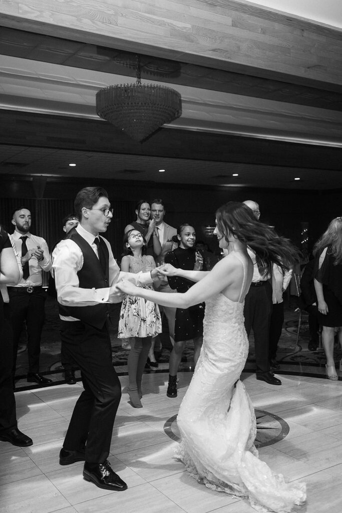A bride and groom dance together in the center of a lively crowd of wedding guests. The brides dress flows as she moves, and onlookers smile and clap, enjoying the celebration in a well-lit reception hall.