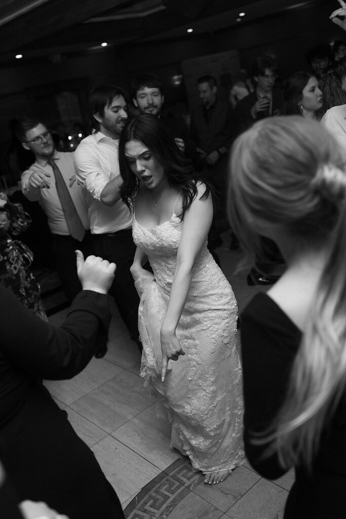 A bride in a lace wedding dress dances energetically on a crowded dance floor, surrounded by guests in formal attire. The atmosphere is lively and joyful. The photo is in black and white.