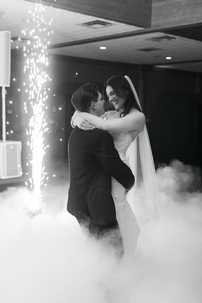 A bride and groom share a joyful dance surrounded by low-lying fog and indoor fireworks, creating a magical atmosphere. The bride wears a veil and smiles at the groom as he holds her. The image is in black and white.