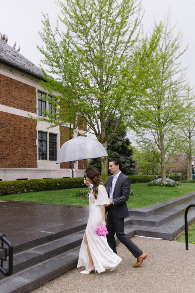 A bride in a white dress holding pink flowers and a transparent umbrella walks with a groom in a suit along a paved path outside a brick building, surrounded by green trees on a rainy day.