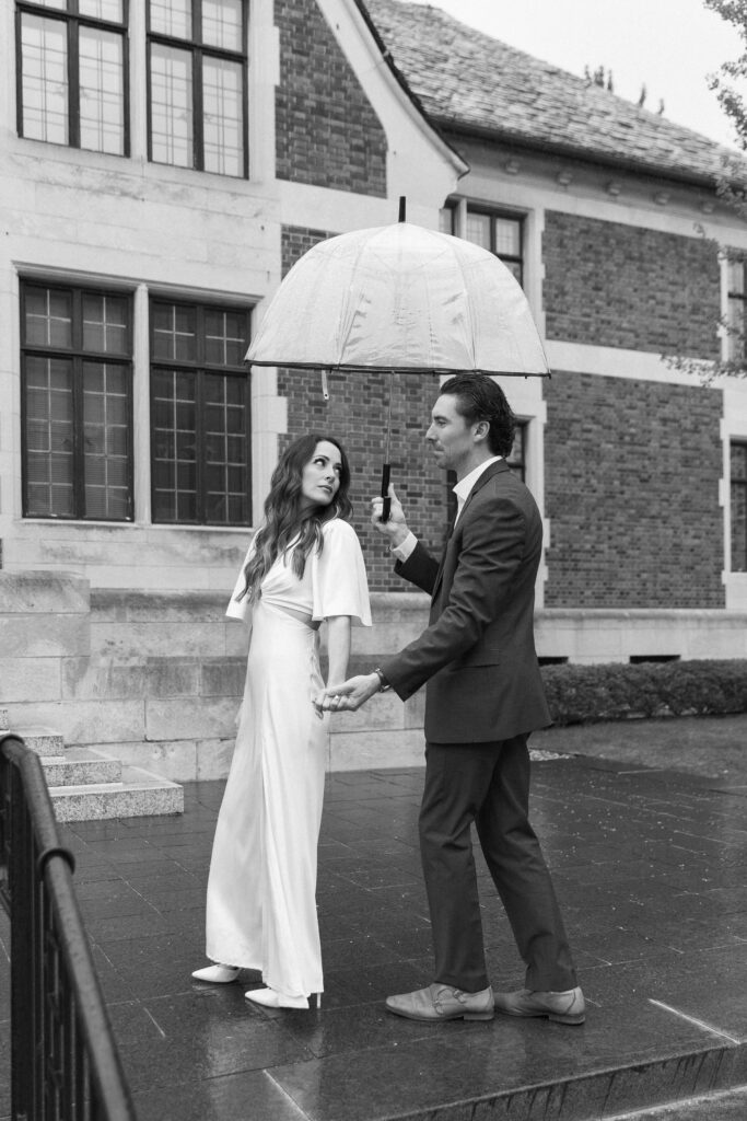 A couple dressed formally stands outside a brick building in the rain. The man in a suit holds an umbrella over the woman in a white dress as they hold hands and look at each other. The photo is in black and white.
