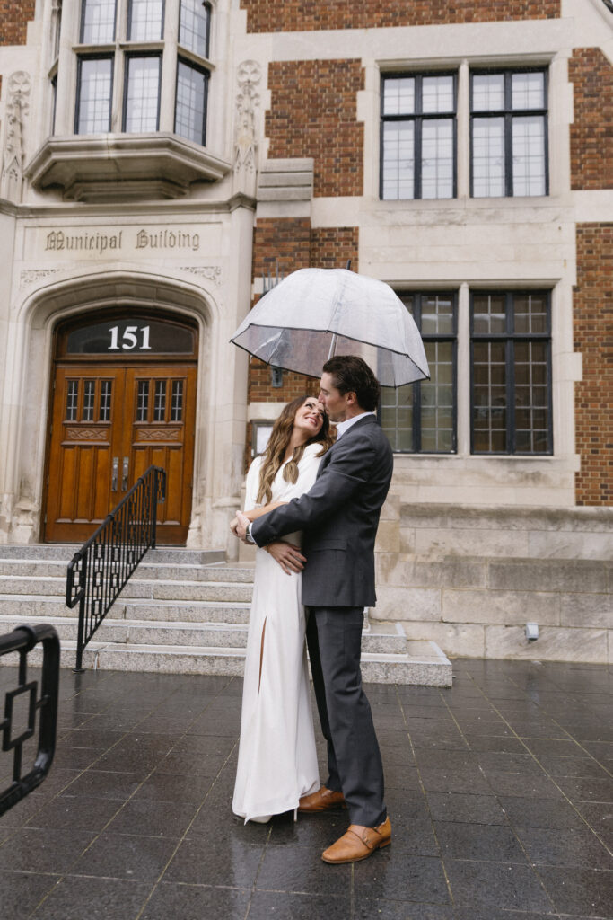 A couple dressed formally embrace under a clear umbrella on a rainy day, standing outside a historic building labeled Municipal Building with the number 151 above wooden doors.