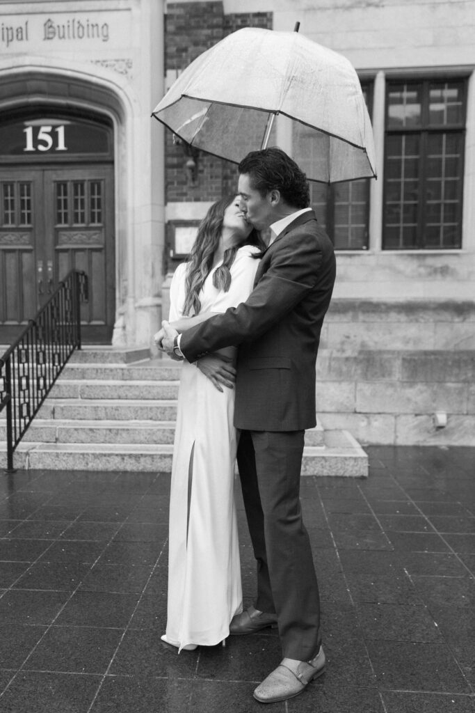 A couple stands in front of a building entrance under a clear umbrella, embracing and sharing a kiss in the rain. The woman wears a long white dress; the man wears a suit. The scene is in black and white.