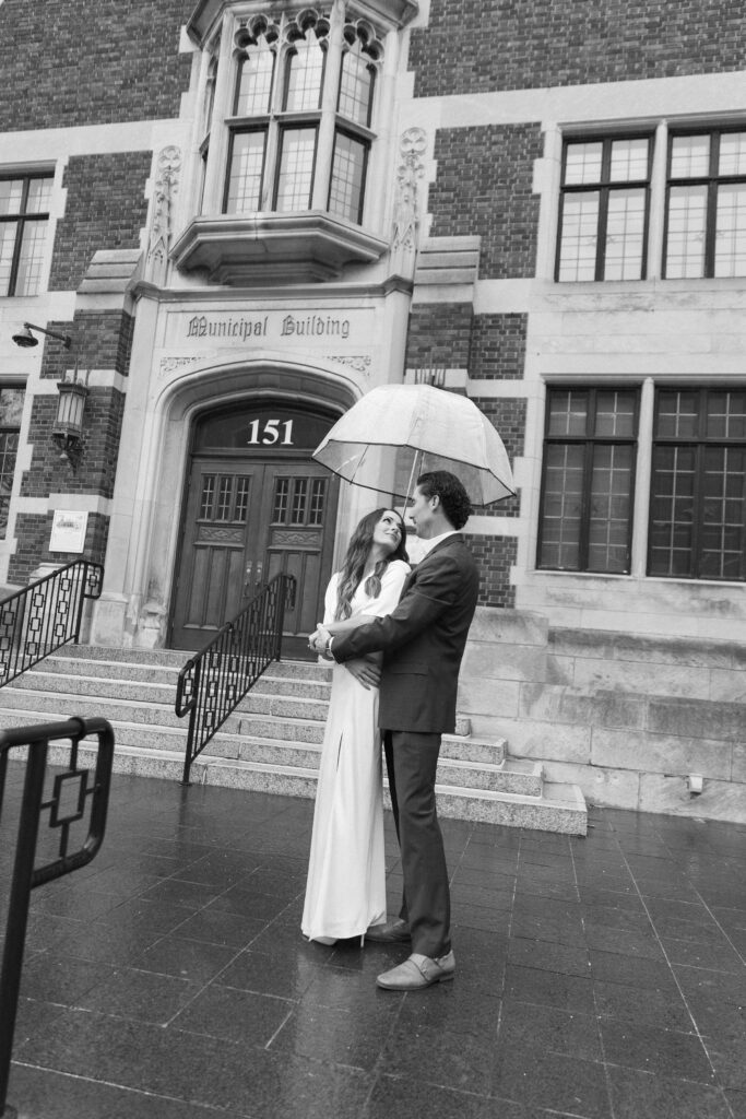 A couple dressed formally stands under an umbrella, gazing at each other in front of a building marked Municipal Building 151. The scene appears romantic and rainy.