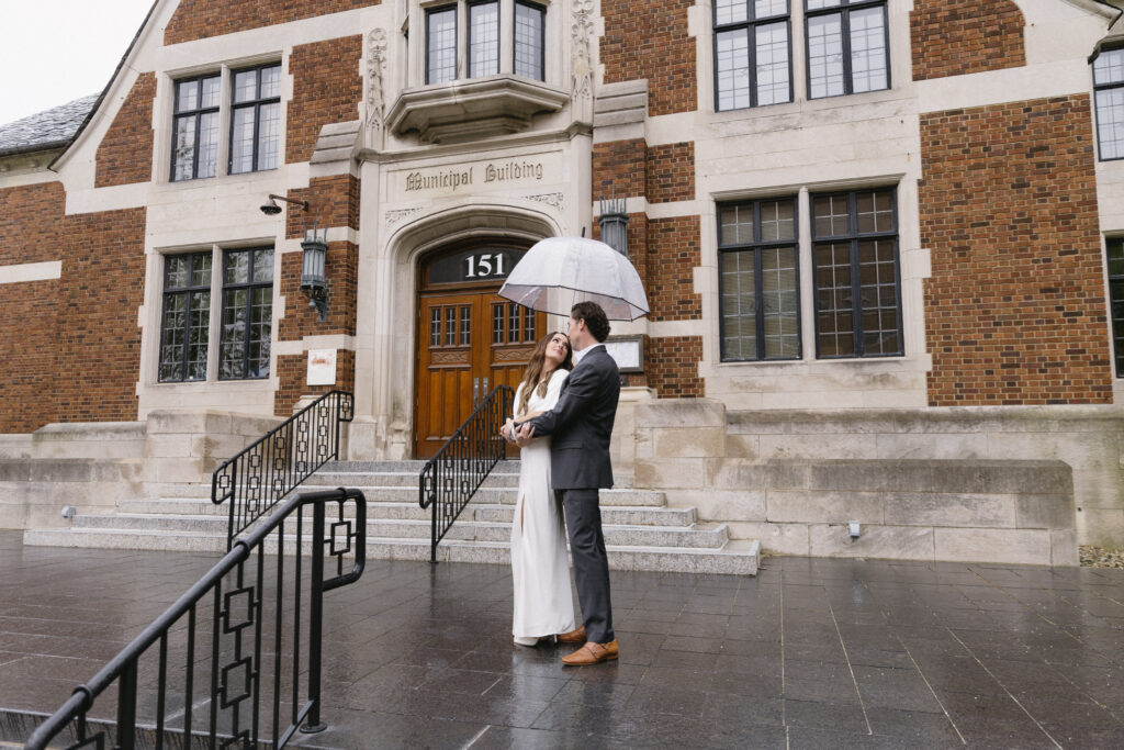A couple dressed for a formal occasion stands under an umbrella on wet steps outside a historic brick building with a sign reading Municipal Building above the entrance.