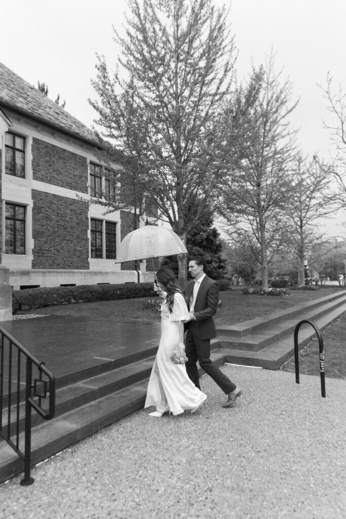 A bride and groom walk outside on a rainy day; the groom holds a clear umbrella over them as they walk up steps near a brick building lined with trees.