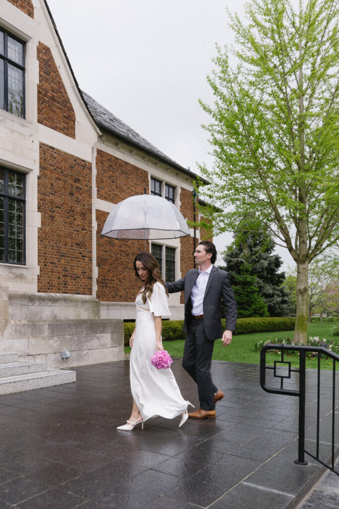 A bride in a white dress holds pink flowers and walks beside a groom in a suit, who carries a clear umbrella. They are outside a brick building with green trees and wet pavement, suggesting recent rain.