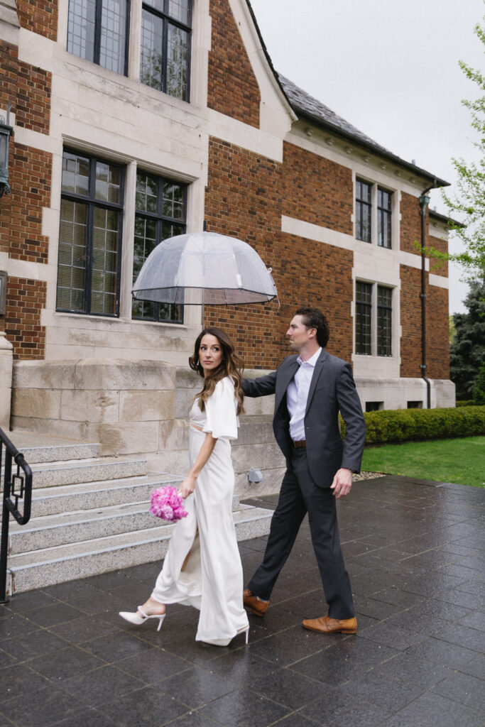 A woman in a white outfit holding pink flowers walks beside a man in a suit who is holding a clear umbrella over them; they are outside a brick building on a wet, overcast day.
