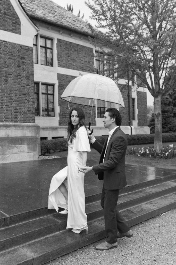 A man in a suit holds an umbrella over a woman in a white dress as she walks up stone steps outside a brick building on a rainy day.