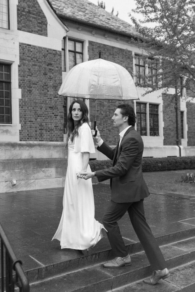 A man in a suit holds an umbrella and helps a woman in a long white dress walk up steps outside a brick building on a rainy day. Both look ahead, and the scene is captured in black and white.