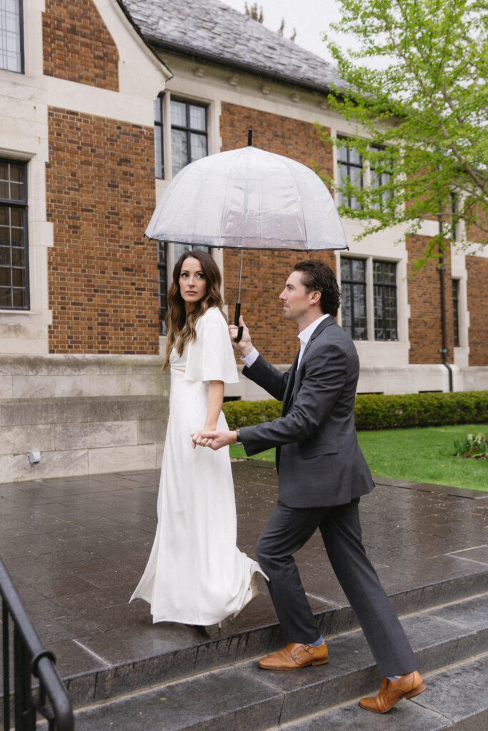 A man in a suit holds an umbrella and leads a woman in a white dress up stone steps outside a brick building on a rainy day. The woman looks at the camera while holding his hand.