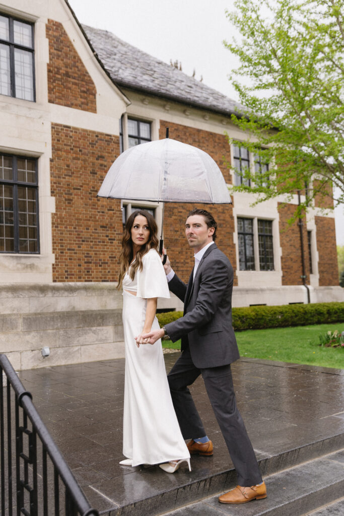 A couple stands outside a brick building on a rainy day. The woman wears a white dress and the man, in a gray suit, holds a transparent umbrella over them as they hold hands and look ahead.