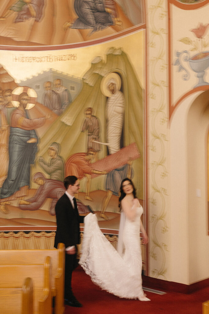 A bride in a white gown walks while a groom in a black suit holds her train, both beside a colorful religious mural inside a church with ornate details and red carpeting.