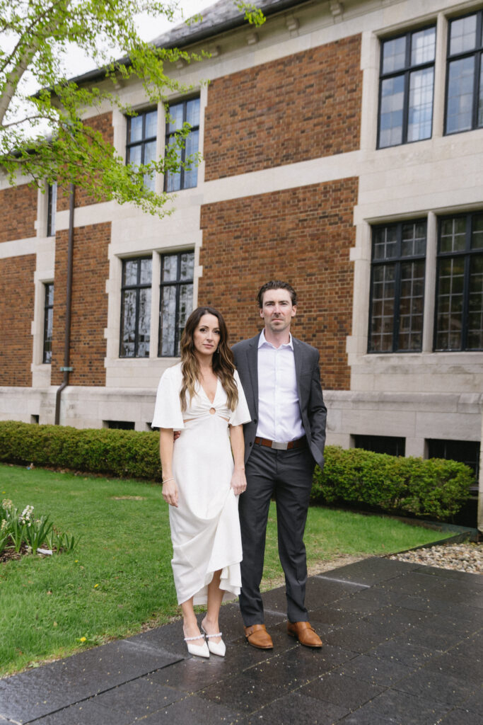 A woman in a white dress and a man in a dark suit stand side by side on a wet path outside a brick building with large windows, surrounded by greenery.