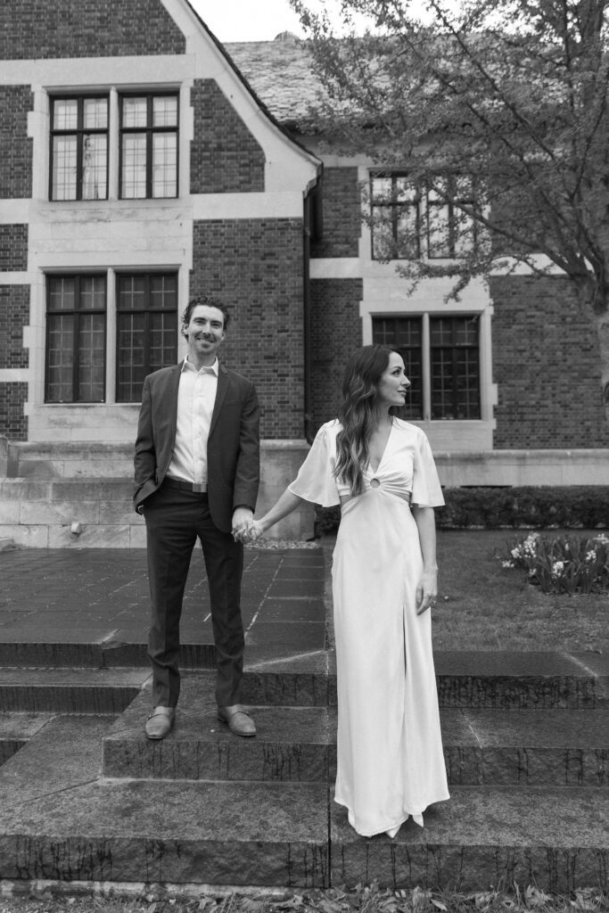 A man in a suit and a woman in a long white dress hold hands while standing on stone steps in front of a large brick building, looking in different directions. The image is in black and white.