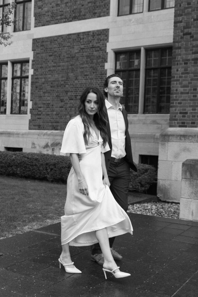 A woman in a white dress and heels walks beside a man in a suit outside a brick building, both looking ahead with serious expressions. The scene is captured in black and white.