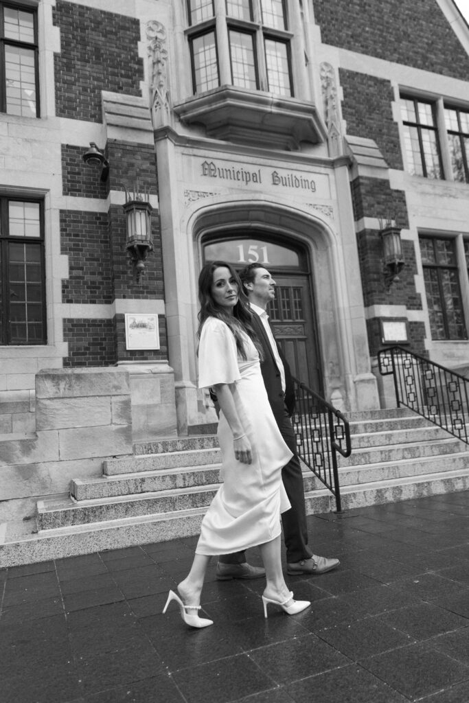 A woman in a white dress and heels walks beside a man in a suit up the steps of a brick Municipal Building. Both appear to be smiling. The image is in black and white.