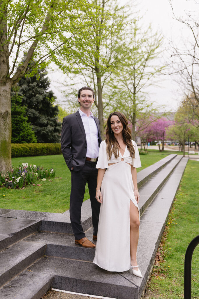 A man in a suit and a woman in a white dress stand together on outdoor stone steps surrounded by green trees and grass, smiling at the camera on a cloudy day.