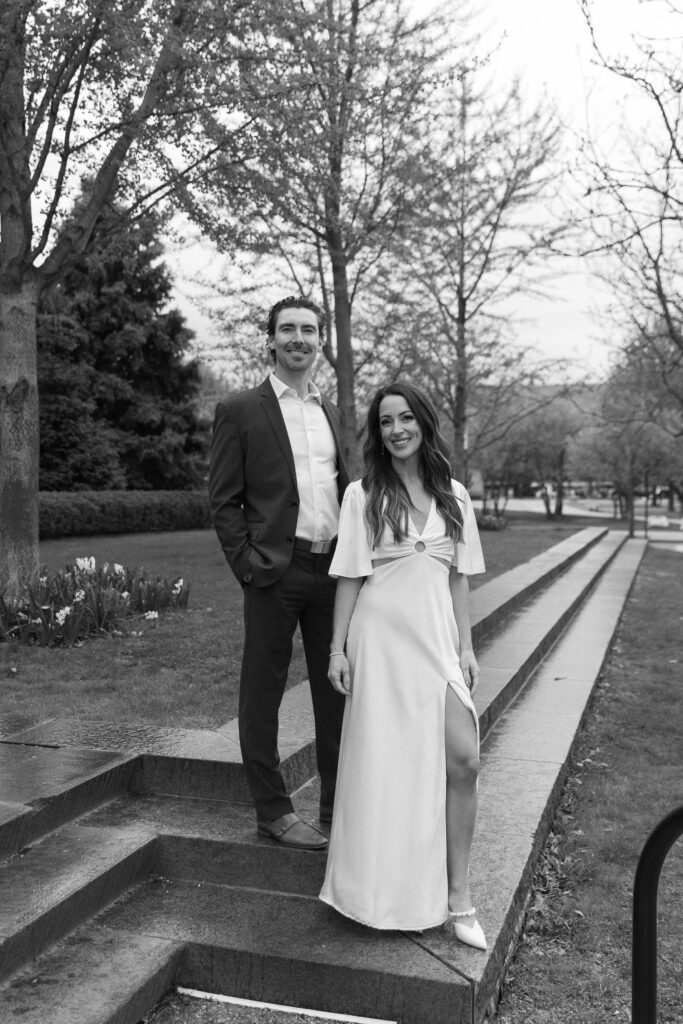 A man in a suit and a woman in a light, flowy dress stand together on outdoor stone steps, surrounded by trees and greenery. Both are smiling, and the setting appears calm and elegant.