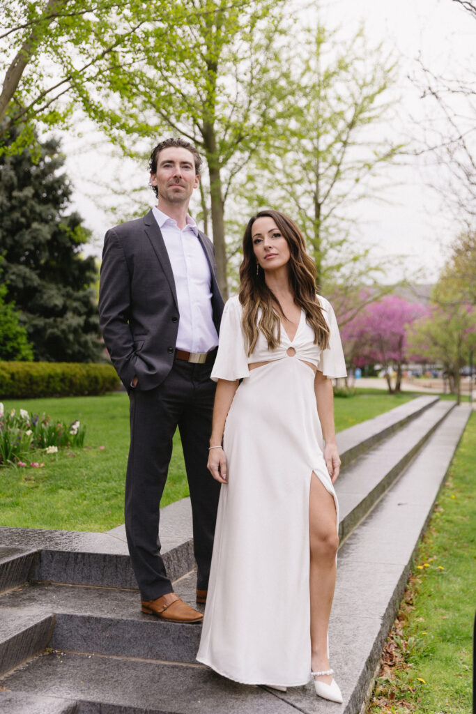 A man in a dark suit and a woman in a white dress stand on stone steps outdoors, surrounded by green grass and leafy trees with spring blossoms. The woman stands slightly ahead, looking toward the camera.